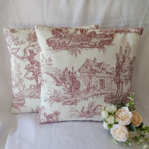 May include: Two cream-colored pillow covers with a red toile pattern featuring a cottage, bridge, and trees. The pattern is reminiscent of a French country design.