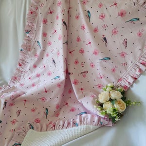 May include: A pink baby blanket with a white ruffle border. The blanket features a repeating pattern of pink flowers, blue birds, and red dragonflies on a white background. The blanket is made of soft cotton and is perfect for cuddling.