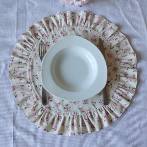 Peut inclure: Une assiette en céramique blanche, un ensemble de fourchette et de couteau sur un set de table floral. Le set de table a un fond crème avec des motifs de roses roses et un bord à volants. La table est sur une nappe blanche texturée.