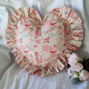 Peut inclure: Un coussin en forme de cœur avec un motif patchwork de roses roses et de motifs floraux. Il a un bord à volants dans un tissu floral assorti. La palette de couleurs comprend le rose, la crème et le bleu clair. Objet décoratif.