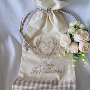 Op de afbeelding: Een witte stoffen tas met een bruine ruitjesrand. De tas heeft een geborduurd windmolenmotief en de tekst "Pain Fait Maison".