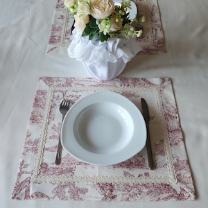Peut inclure: Un bol en céramique blanche avec une bordure en dentelle blanche repose sur un set de table à motifs floraux rouge et blanc. Un vase en céramique blanche avec un arrangement floral se trouve sur un set de table similaire à l'arrière-plan. Deux fourchettes et un couteau sont placés sur le set de table.