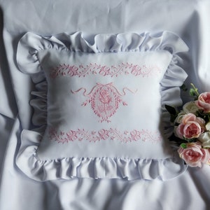 Pode incluir: Uma almofada branca com uma borda franjada e um desenho floral bordado em rosa. A almofada está sobre um fundo branco.