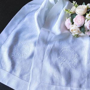 Peut inclure: Chemin de table en lin blanc avec broderies de couronnes florales. Le chemin de table présente une bordure délicate à points de feston et est drapé sur une surface sombre. Un bouquet de fleurs roses et blanches repose sur le chemin.