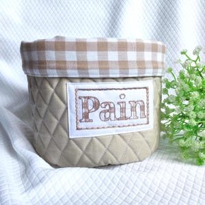 Peut inclure: Un panier en tissu beige matelassé avec une bordure à carreaux vichy. Le mot "Pain" est brodé sur un patch rectangulaire blanc à l'avant. Le panier est cylindrique et repose sur une surface blanche.