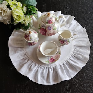 Peut inclure: Un dessous de plat rond en lin blanc avec des volants, sur lequel sont posés une théière, un pot à crème, un sucrier et une tasse à thé à motifs floraux. L'ensemble à thé est décoré de roses roses, violettes et blanches.
