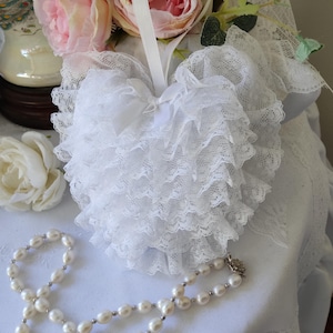 May include: White lace heart-shaped pillow with a white satin bow. The pillow is layered with ruffles and has a loop for hanging.