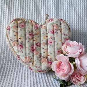 Op de afbeelding: Hartvormige, gewatteerde pannenlap met een bloemenpatroon in roze, geel en crème. De pannenlap heeft een lus om op te hangen. Een boeket roze rozen ligt ernaast.