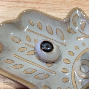 May include: A ceramic dish with a hand-shaped design features a white and brown eye-shaped stone. The dish has a light blue glaze with a beige pattern.