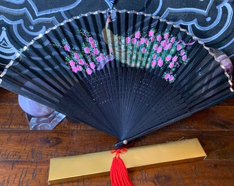 May include: A black folding fan with a silver trim and pink floral design. The fan is closed and has a red tassel.