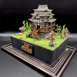 Peut inclure: Maquette détaillée d'un château japonais à plusieurs niveaux, avec des accents en bois marron et un toit gris. Le modèle repose sur une base herbeuse verte avec des arbres et des figurines miniatures. Le logo "Age of Empires" est visible.