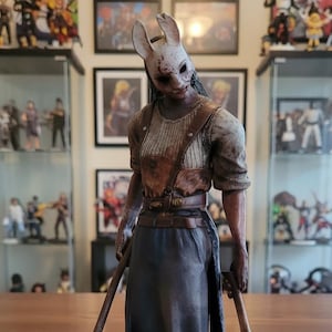 Op de afbeelding: Gedetailleerd beeldje van een personage uit Dead by Daylight, The Huntress, staat op een zwarte basis. Het beeldje draagt een wit konijnenmasker, een bruin schort en houdt twee bebloede bijlen vast. Het beeldje staat op een houten tafel.