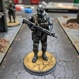 May include: A miniature figurine of a soldier in a gas mask and tactical gear, holding a rifle. The figure is painted in shades of black, brown, and gray, standing on a circular base. Another similar figure is visible in the background.