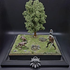 Può includere: Diorama in miniatura raffigurante una scena di The Witcher. Include una figura con una spada, due figure di mostri, un albero e un medaglione a forma di lupo. La base è quadrata con cornice nera, su sfondo scuro.