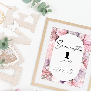 May include: A framed print with a watercolor floral design in shades of pink and purple. The print features a white circle with the text "Samantha 1 year old 21/05/21".