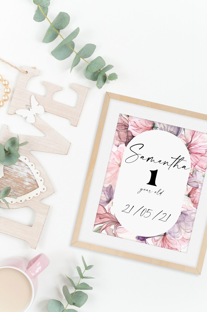 May include: A framed print with a watercolor floral design in shades of pink and purple. The print features a white circle with the text "Samantha 1 year old 21/05/21".