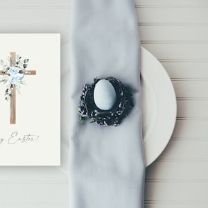 May include: A white card with a wooden cross decorated with blue flowers and greenery. The text "Happy Easter!" is written below the cross. A white plate with a blue egg in a nest is on a light blue napkin.