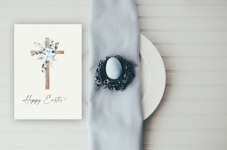 May include: A white card with a wooden cross decorated with blue flowers and greenery. The text "Happy Easter!" is written below the cross. A white plate with a blue egg in a nest is on a light blue napkin.