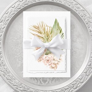 May include: Two white cards with a watercolor illustration of tropical leaves and blush pink roses tied together with a white satin ribbon. The cards are on a silver tray with a decorative border.