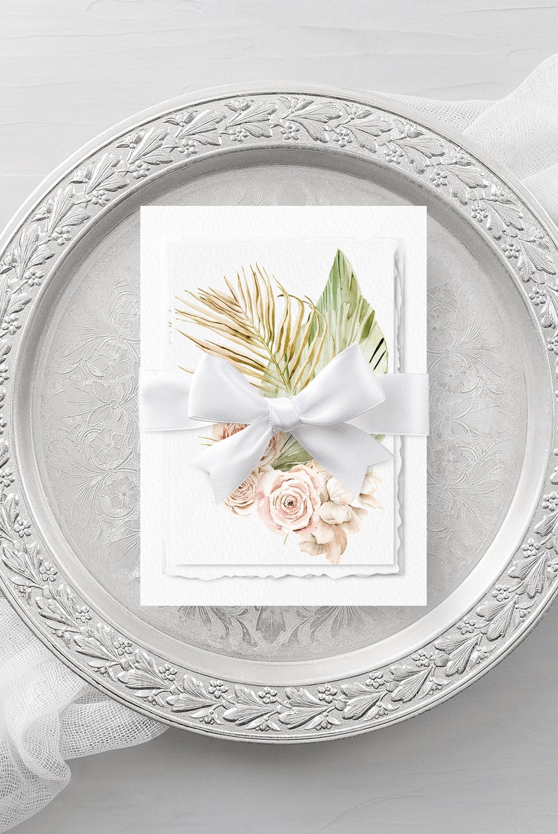 May include: Two white cards with a watercolor illustration of tropical leaves and blush pink roses tied together with a white satin ribbon. The cards are on a silver tray with a decorative border.