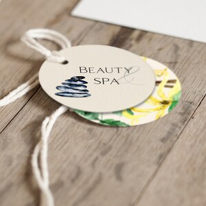 May include: A round, cream-colored tag with the text "Beauty & Spa" and a stack of gray stones. The tag is attached to a white string.