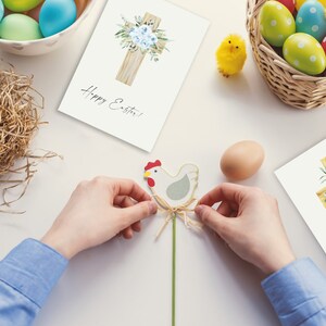 May include: Two white cards with a watercolor illustration of a wooden cross with blue flowers and the text "Happy Easter!" on each card. A person is holding a wooden chicken figurine with a green stem and a brown twine bow.