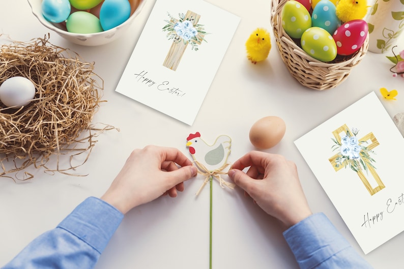May include: Two white cards with a watercolor illustration of a wooden cross with blue flowers and the text "Happy Easter!" on each card. A person is holding a wooden chicken figurine with a green stem and a brown twine bow.