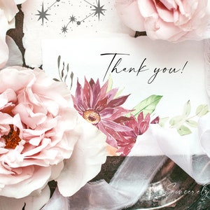 May include: A white card with the text "Thank you!" written in black cursive. The card is decorated with watercolor flowers in shades of pink and green. The card is surrounded by pink roses and white lace.