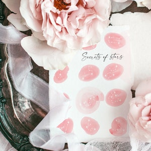 May include: A white card with pink watercolor circles featuring constellations and the text "Secrets of stars". The card is surrounded by pink flowers and white fabric.
