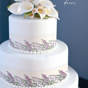 May include: A three-tiered white cake decorated with a floral arrangement of white calla lilies and other flowers. The cake has a band of light beige with a floral design of purple flowers and green leaves. The text "Cake decor" is written on the right side.
