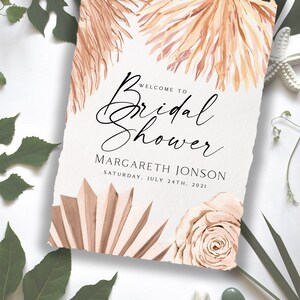 May include: A white card with a torn edge, featuring a watercolor illustration of dried palm leaves and a rose. The text "Welcome to Bridal Shower" is written in black cursive script. Below the text is the name "MARGARETH JONSON" and the date "SATURDAY, JULY 24TH, 2021".