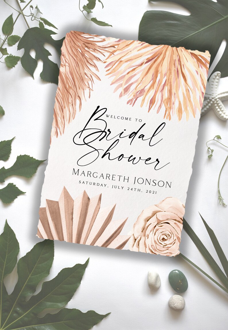 May include: A white card with a torn edge, featuring a watercolor illustration of dried palm leaves and a rose. The text "Welcome to Bridal Shower" is written in black cursive script. Below the text is the name "MARGARETH JONSON" and the date "SATURDAY, JULY 24TH, 2021".