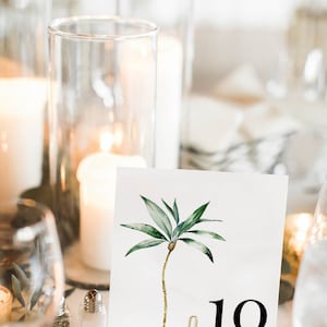May include: A white card with a watercolor illustration of a palm tree and the number 10 printed in black. The card is propped up on a gold metal stand.