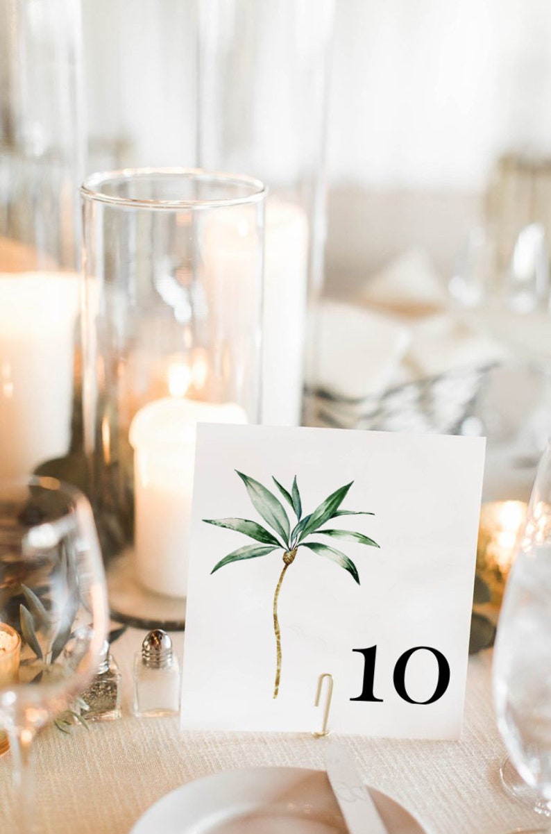 May include: A white card with a watercolor illustration of a palm tree and the number 10 printed in black. The card is propped up on a gold metal stand.