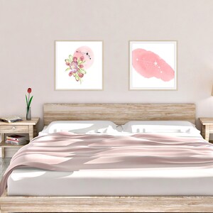 May include: Two framed watercolor prints on a white wall above a bed with a pink bedspread. The print on the left features a pink watercolor circle with a floral design and a constellation. The print on the right features a pink watercolor shape with a constellation.