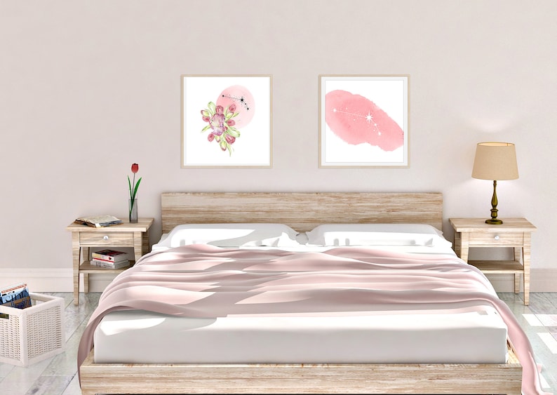 May include: Two framed watercolor prints on a white wall above a bed with a pink bedspread. The print on the left features a pink watercolor circle with a floral design and a constellation. The print on the right features a pink watercolor shape with a constellation.