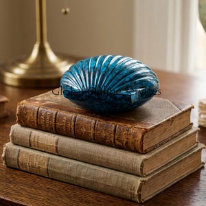 May include: A blue, shell-shaped ornament with white detailing rests on a pile of old books. The books have worn, textured covers in shades of brown and beige. The shell has a shiny finish and is placed on the top book.