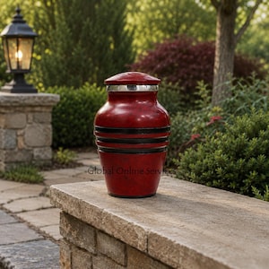 May include: A red cremation urn with a mottled finish, featuring a silver-toned lid and two prominent black horizontal bands encircling the midsection. The urn has a tapered shape with a wide body and a narrower base, displaying a smooth texture.