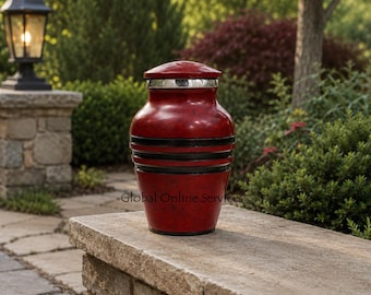 Op de afbeelding: Een rode urn met een gemarmerde afwerking, voorzien van een zilverkleurig deksel en twee opvallende zwarte horizontale banden rond het middengedeelte. De urn heeft een taps toelopende vorm met een brede romp en een smallere basis.