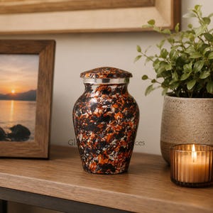 May include: A cremation urn featuring a mottled, abstract pattern of black, orange, and white colors. The urn has a rounded body, a narrow neck, and a matching lid with a silver-toned metal band separating the lid from the main body of the vessel.