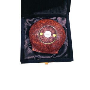Op de afbeelding: Een rond, roodbruin object met een goudkleurige ring en een wit centrum, gepresenteerd in een zwarte fluwelen doos. De tekst "Glob line" is zichtbaar op het object.
