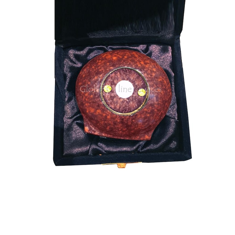 Op de afbeelding: Een rond, roodbruin object met een goudkleurige ring en een wit centrum, gepresenteerd in een zwarte fluwelen doos. De tekst "Glob line" is zichtbaar op het object.