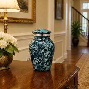 May include: A cremation urn featuring a mottled teal and dark blue finish with a metallic silver band around the lid. The vessel has a rounded body and a secure, matching lid, displaying a textured, marbled appearance across its entire surface.
