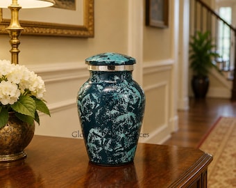 May include: A cremation urn featuring a mottled teal and dark blue finish with a metallic silver band around the lid. The vessel has a rounded body and a secure, matching lid, displaying a textured, marbled appearance across its entire surface.