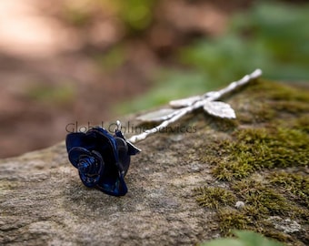 May include: A dark blue rose sculpture with a silver stem and leaves rests on a mossy rock. The rose's petals are intricately detailed, and the silver stem has a textured appearance. The background is blurred, suggesting an outdoor setting.