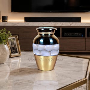 Puede incluir: Una urna decorativa con una base de oro metálico y una tapa dorada a juego. La sección central está adornada con una banda de piezas de nácar blanco de forma hexagonal. El recipiente tiene un acabado liso y pulido con una forma cónica clásica.