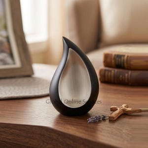 May include: A black and silver teardrop-shaped urn. The urn has a sleek, modern design with a black exterior and a silver interior. A wooden cross and lavender sprig are next to the urn. The text "Global Online Services" is printed on the urn.
