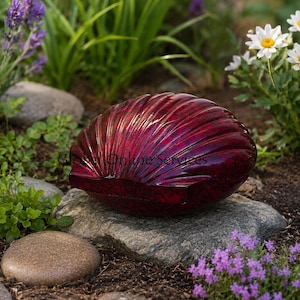 May include: A glossy, deep red decorative shell-shaped object with a textured surface. The object is resting on a grey stone, surrounded by green plants, purple flowers, and white daisies. The shell has a reflective surface.