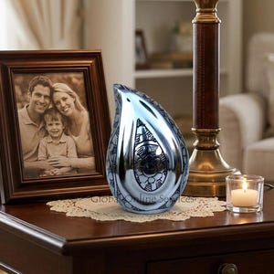 Könnte beinhalten: Eine polierte, silberne Urne in Tropfenform mit aufwendigen floralen und geometrischen Mustern. Die Urne steht auf einem Holztisch neben einem gerahmten Familienfoto, einer brennenden Kerze und einer Lampe.