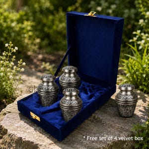 May include: Four silver urns with detailed floral patterns, displayed in a deep blue velvet box with a gold clasp. The box is placed on a stone surface. The text at the bottom says "Free set of 4 velvet box".